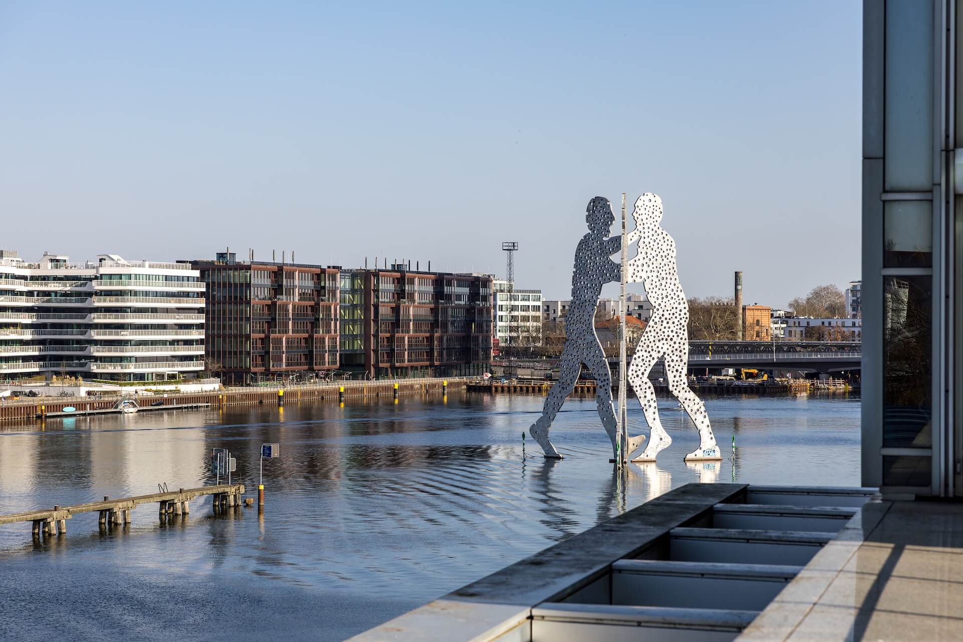 Skulptur „Molecule Man“ im Wasser der Spree vor moderner Uferbebauung in Berlin bei klarem Himmel.