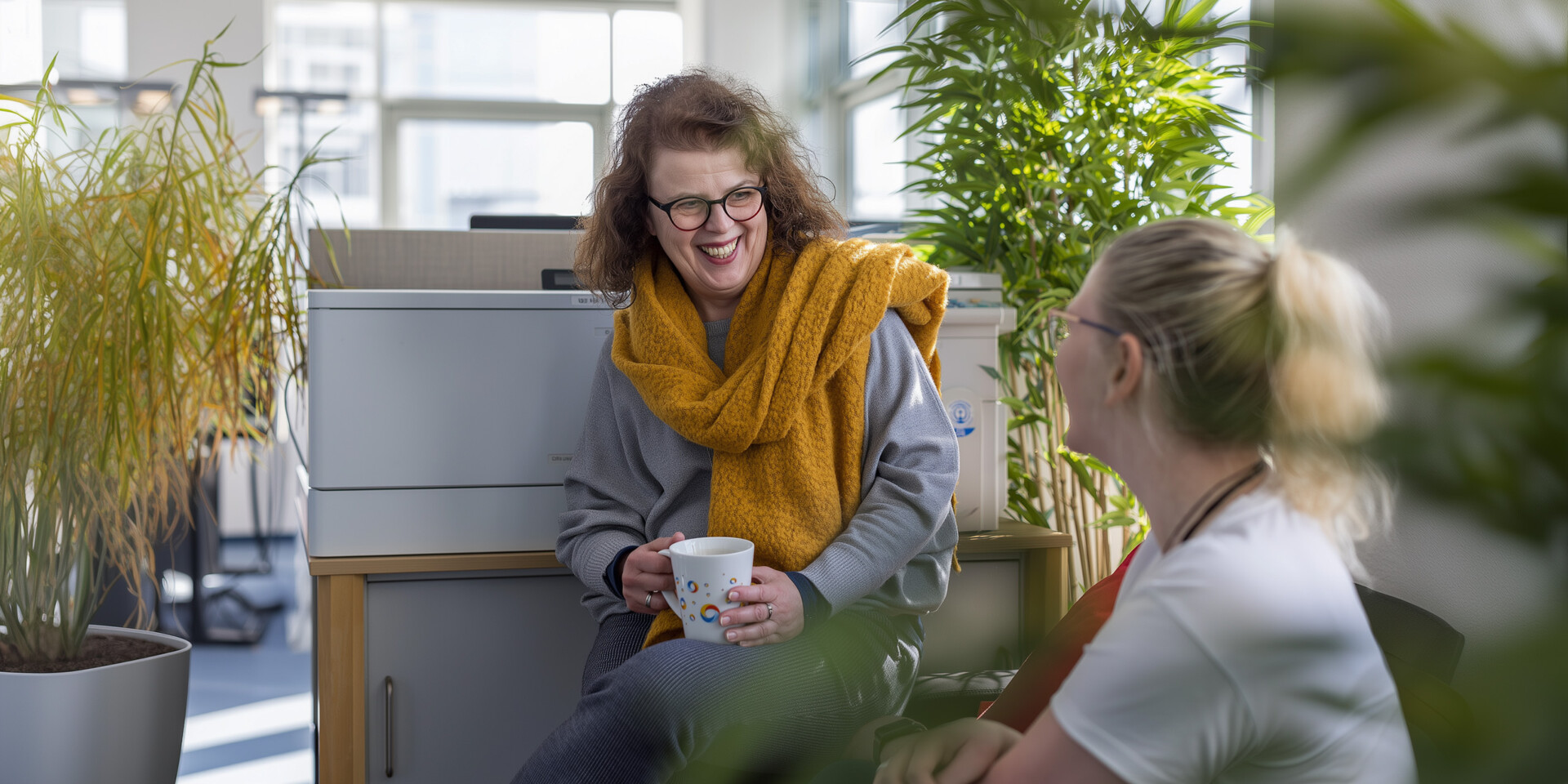 Zwei Reiseprofis unterhalten sich lachend im begrünten Büro, eine Person mit gelbem Schal hält eine Tasse